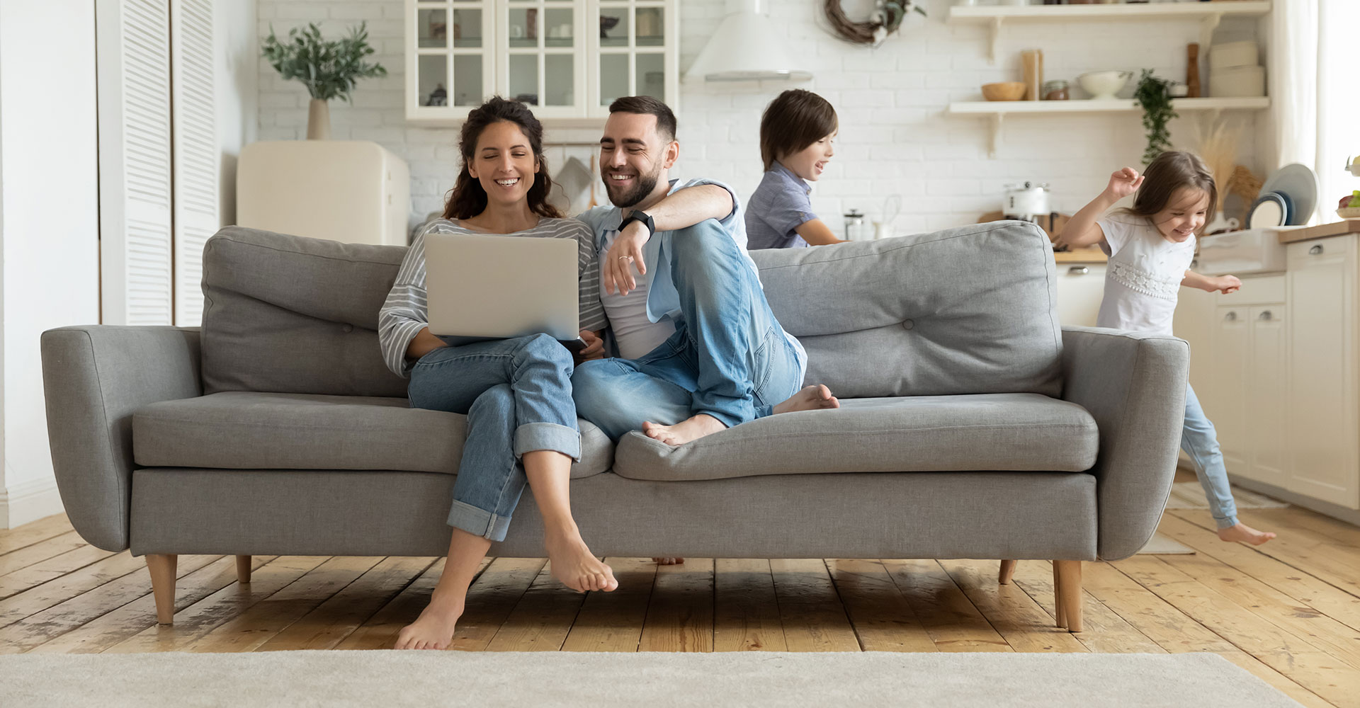 Family of four relaxing together on gray couch in bright living room with open kitchen in background.