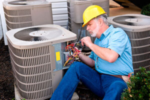 Skilled HVAC technician in yellow hard hat and blue shirt performing maintenance on outdoor air conditioning unit with wiring visible.