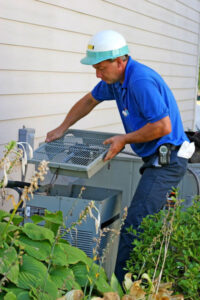 Professional HVAC technician in blue uniform and safety helmet inspecting and cleaning air conditioning unit filter outdoors.