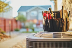 Tool bag filled with wrenches and equipment sits on an air conditioning unit in front of a house with red brick fence.