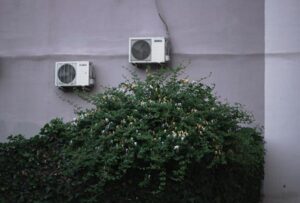 Two white air conditioning units mounted on a gray wall above green climbing ivy with white flowers.