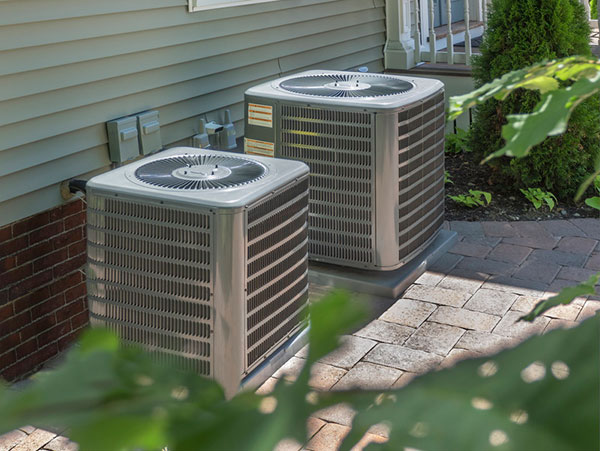 Two large HVAC air conditioning condenser units installed outside a brick home with landscaping nearby.